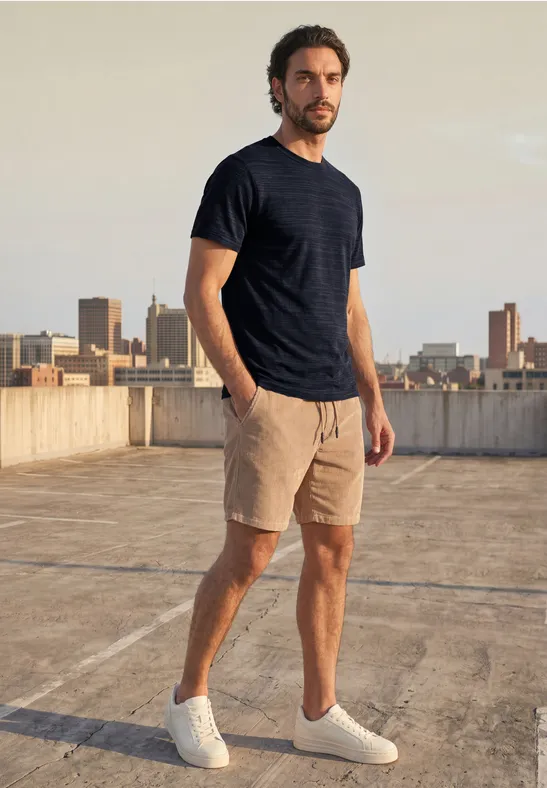 Man in donkerblauw T-shirt, beige korte broek en witte sneakers op een dak met stadsachtergrond.