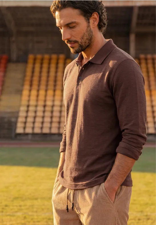 Ein Mann in braunem Langarm-Poloshirt steht mit den Händen in den Taschen auf einem Stadionrasen.