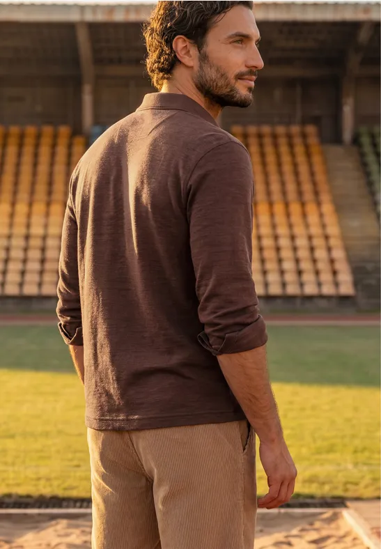 Ein Mann im braunen Outfit steht auf der Sportanlage vor leeren Tribünen und blickt über die Schulter.