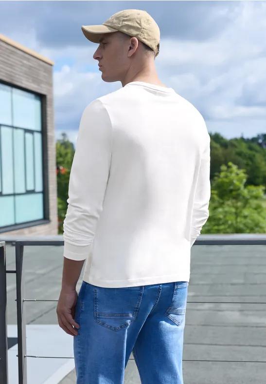 Ein Mann steht auf einer Terrasse, trägt ein weißes Langarmshirt, blaue Jeans und eine Kappe.