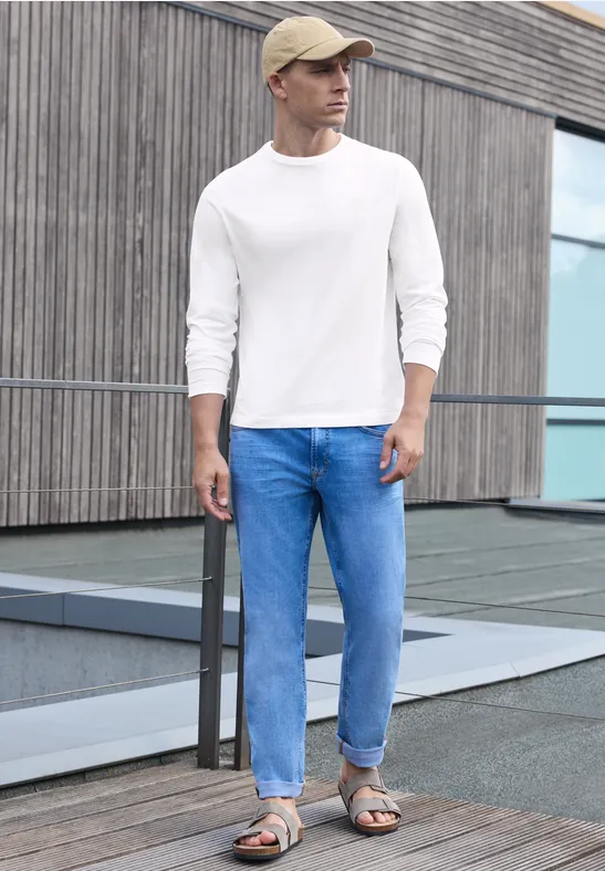 Ein Mann in Jeans, weißem Pullover und Sandalen lehnt an einem Geländer vor einem modernen Gebäude.