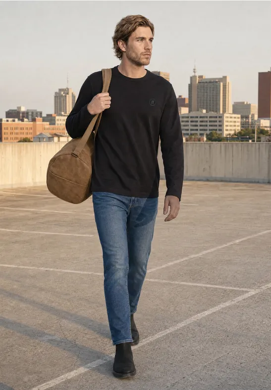 Mann in Jeans und Pullover trägt Reisetasche auf Parkdeck vor städtischer Skyline.