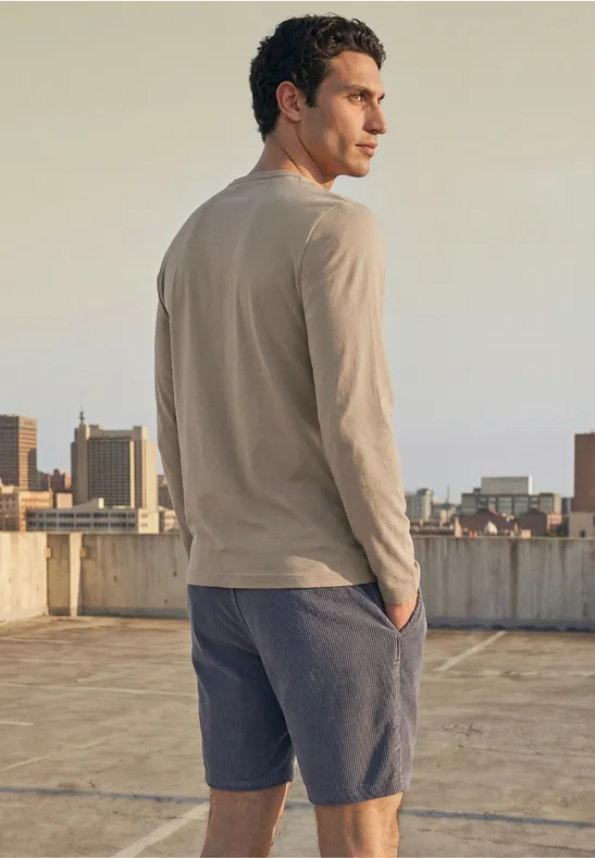 Ein Mann steht auf einem Dach und blickt über die Stadt, trägt ein beiges Langarmshirt und blaue Shorts.