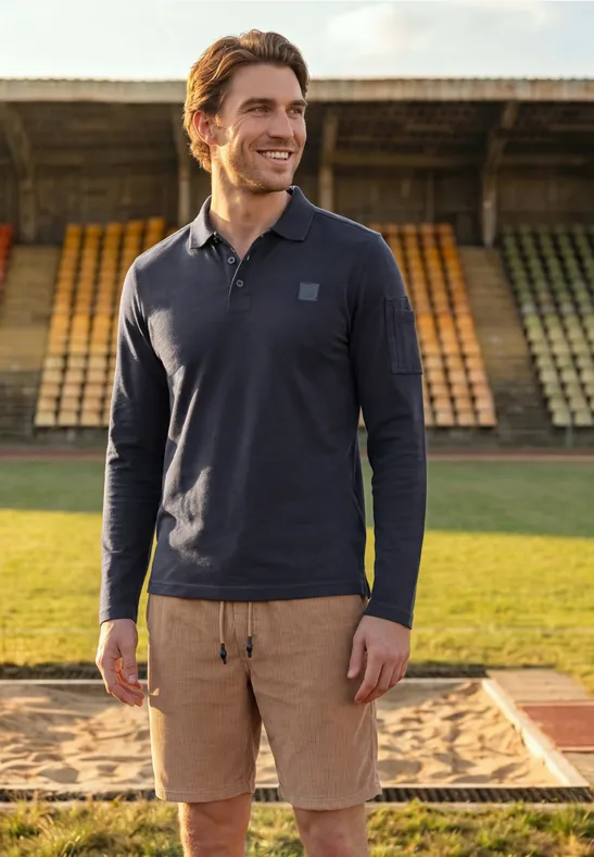 Man in donkerblauw poloshirt met lange mouwen en beige short glimlacht op een atletiekbaan voor lege tribunes.