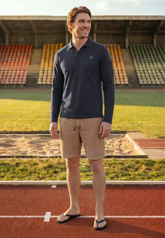 Een man in poloshirt, cargoshort en slippers staat glimlachend op de atletiekbaan voor kleurrijke tribunes.