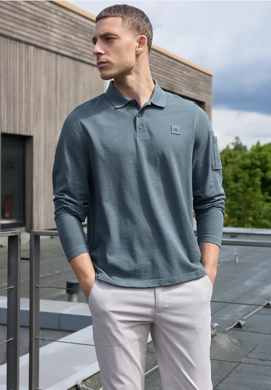 Ein Mann steht auf einem Balkon, trägt ein langärmeliges grünes Polohemd und helle Hose, blickt zur Seite.