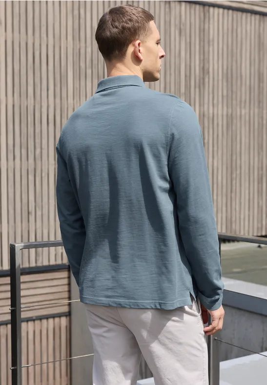 Ein Mann steht von hinten mit einem petrolfarbenen Langarm-Poloshirt und hellen Hosen auf einem Balkon.