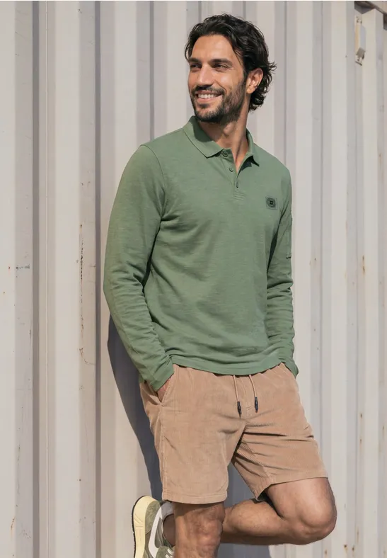 Mann lehnt lächelnd in grüner Poloshirt und beigen Shorts vor einer Metallwand.