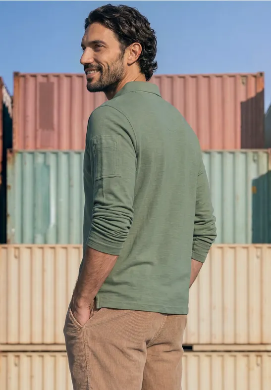 Ein bärtiger Mann in grünem Langarmshirt und brauner Hose blickt über die Schulter vor bunten Frachtcontainern.