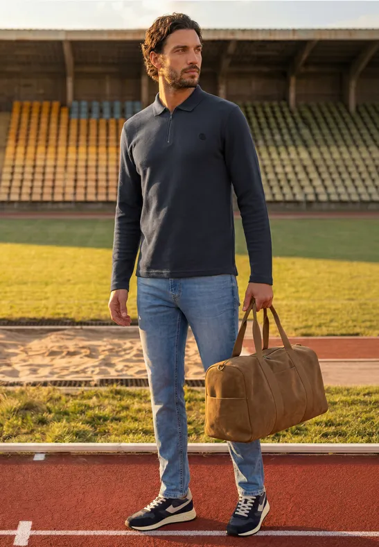 Man met pet, blauw langarm-poloshirt, spijkerbroek en sneakers, bruine tas op atletiekbaan