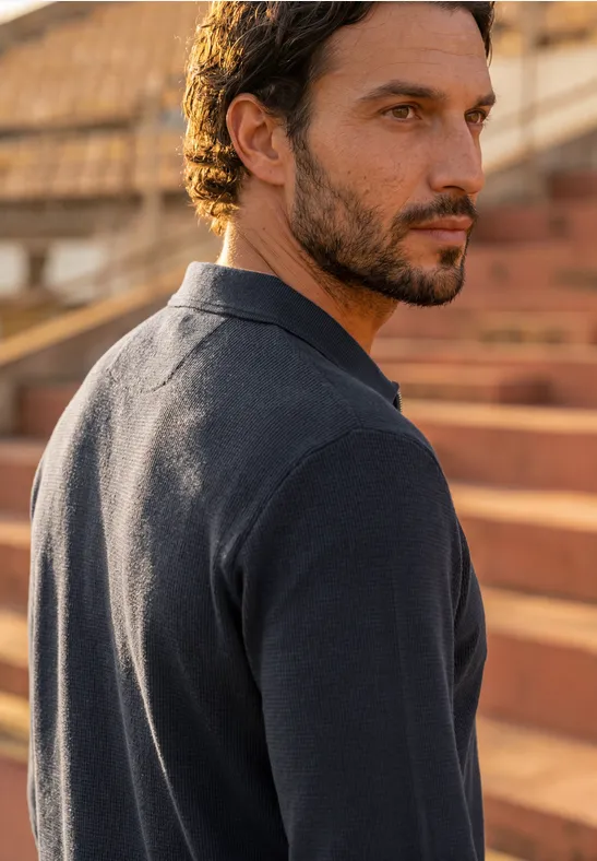 Man met donker haar en baard draagt een donkerblauwe polo en zit op buitentribunes.