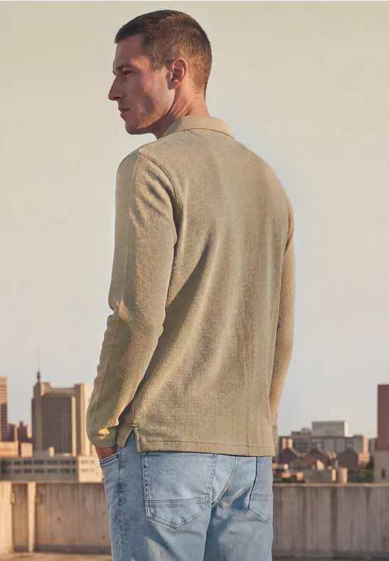 Mann im hellen Pullover und Jeans, der auf einem Dach mit Stadtkulisse steht.