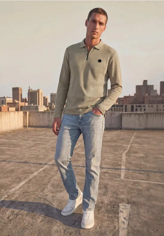Ein Mann auf einem Dach mit hellgrünem Poloshirt, Jeans und weißen Sneakern vor einer Stadtansicht.