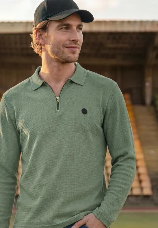 Ein Mann mit grünem Langarm-Poloshirt und schwarzer Baseballkappe steht vor einer Tribüne.