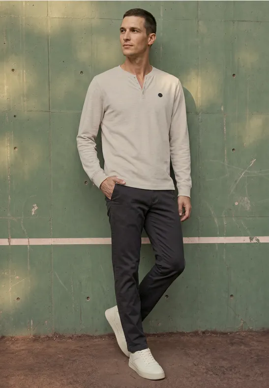 Ein Mann im hellen Pullover, dunkler Hose und weißen Sneakern steht vor einer grünen Wand.