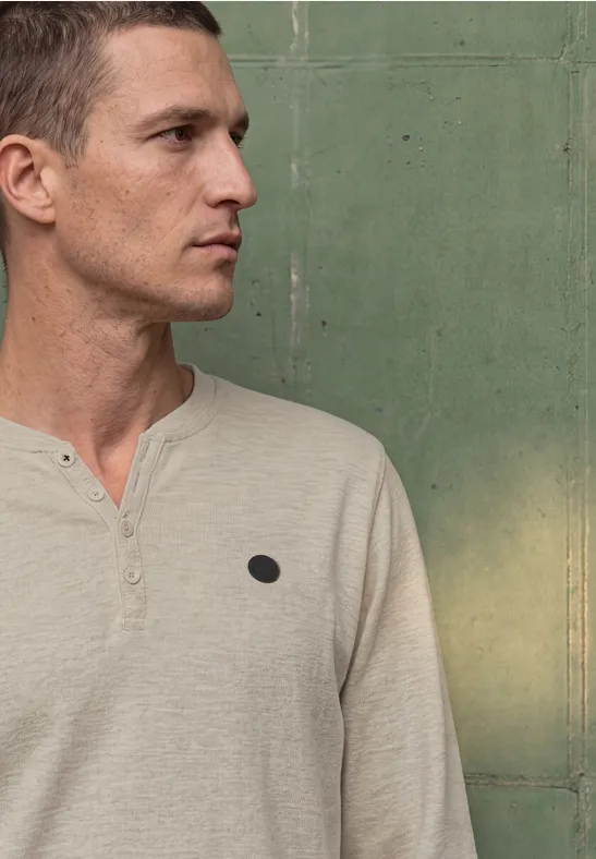 Ein Mann im Profil trägt ein hellbeiges, langärmeliges Shirt mit Knöpfen vor grünem Hintergrund.