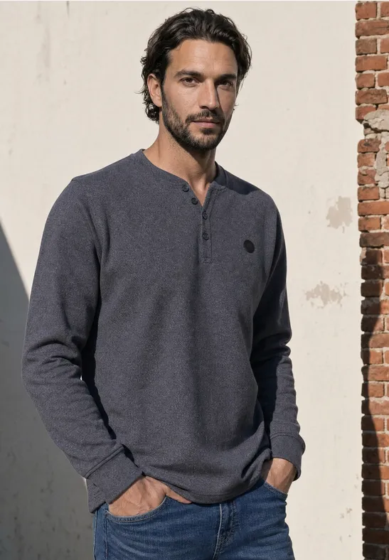 Ein Mann lehnt an einer Mauer und trägt ein dunkelblaues Henley-Shirt.