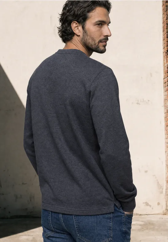 Ein Mann steht von hinten vor einer hellen Wand, trägt grauen Pullover und Jeans im weichen Licht.
