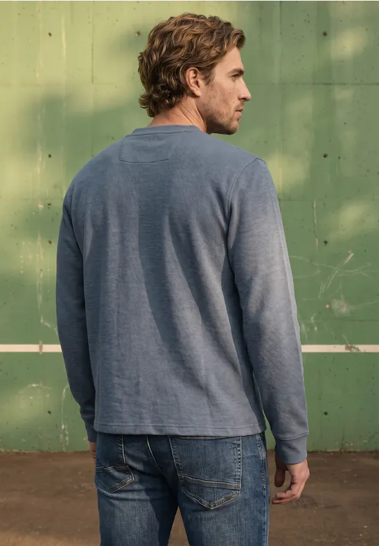 Ein Mann steht von hinten in Jeans und langärmeligem Shirt vor einer grünen Wand.