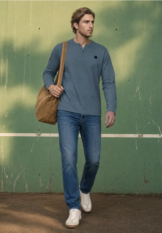 Ein Mann in Jeans, blauem Longsleeve-Shirt und weißen Sneakern trägt eine braune Schultertasche vor grünem Hintergrund.