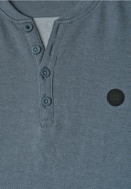 Nahaufnahme eines graublauen Henley-Shirts mit Knopfleiste und schwarzem runden Logo-Patch.