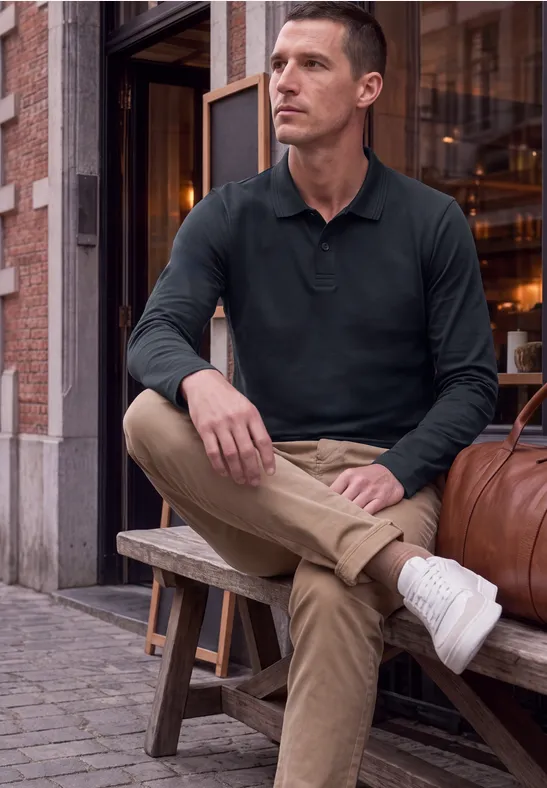 Mann in lässigem Outfit (Polo, Chino, Sneakers) sitzt entspannt auf einer Holzbank vor urbanem Gebäude.