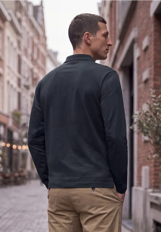 Ein Mann in schwarzem Pullover und beigen Hosen steht in einer gepflasterten Gasse.