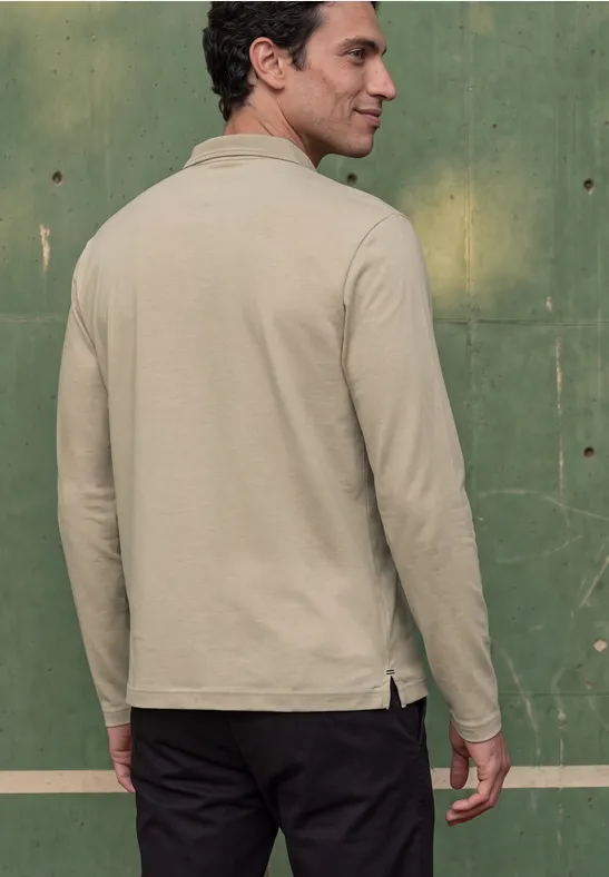 Ein Mann in beiger Langarmjacke steht vor einer grünen Wand, Rücken zur Kamera.