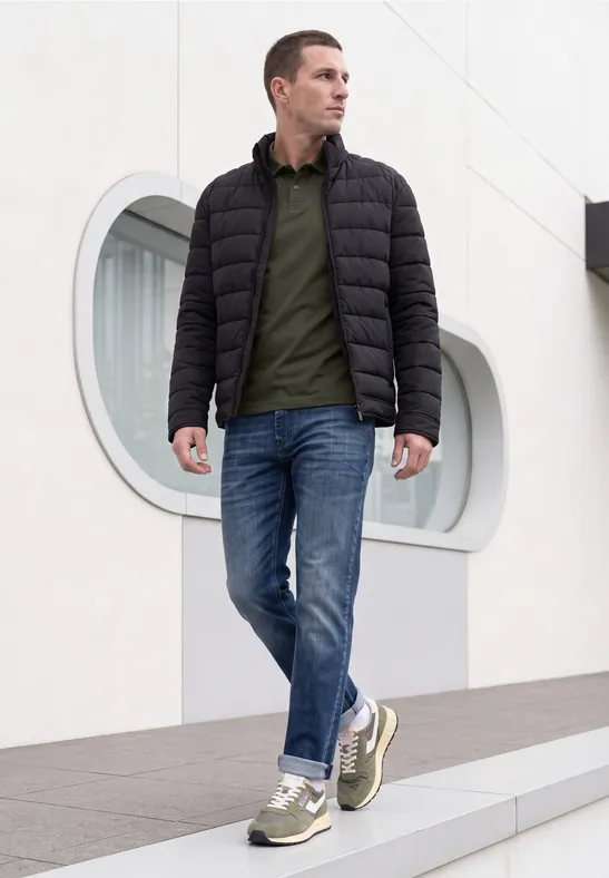 Ein Mann in dunkel gefütterter Jacke, grünem Poloshirt, Jeans und Sneakern spaziert vor modernem Gebäude.