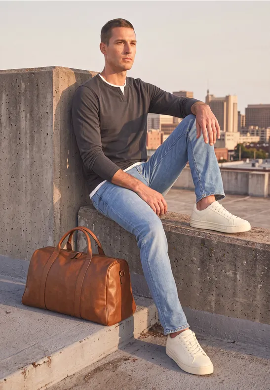 Junger Mann in Jeans und Pullover sitzt mit Lederreisetasche auf einer Betonmauer und blickt auf die Stadt.