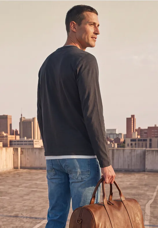 Ein Mann im Langarmshirt und Jeans steht auf einem Dach und hält eine Ledertasche, Blick zur Stadt gerichtet.