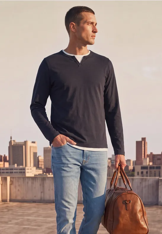 Ein Mann steht auf einem Dach mit Stadtblick, trägt Jeans, schwarzen Pullover und hält eine Ledertasche.