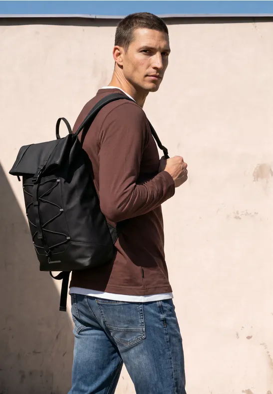 Ein Mann mit braunem Pullover und Jeans trägt einen schwarzen Rucksack vor einer hellen Wand.