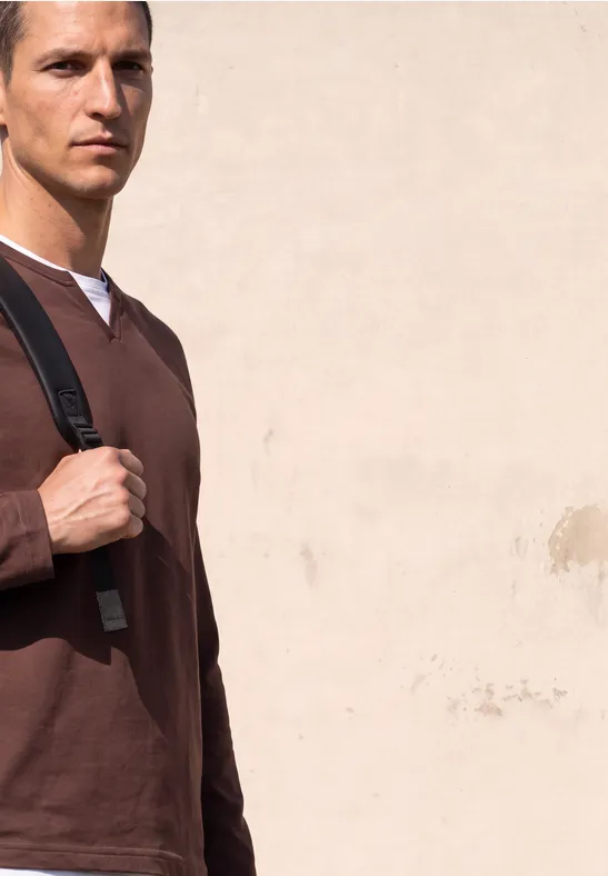Ein junger Mann in braunem Pullover mit Rucksack vor einer hellen Wand.