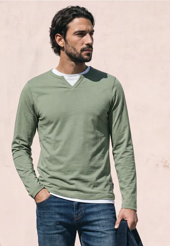 Ein Mann in grünem Langarmshirt und Jeans steht vor einer hellen Wand und blickt seitlich.