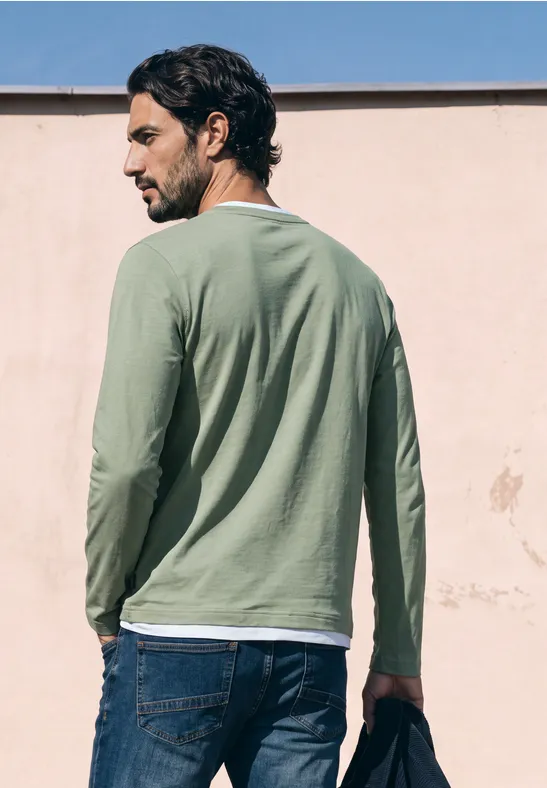 Ein Mann steht mit dem Rücken zur Kamera, trägt grünes Langarmshirt und Jeans, hält eine Sonnenbrille.