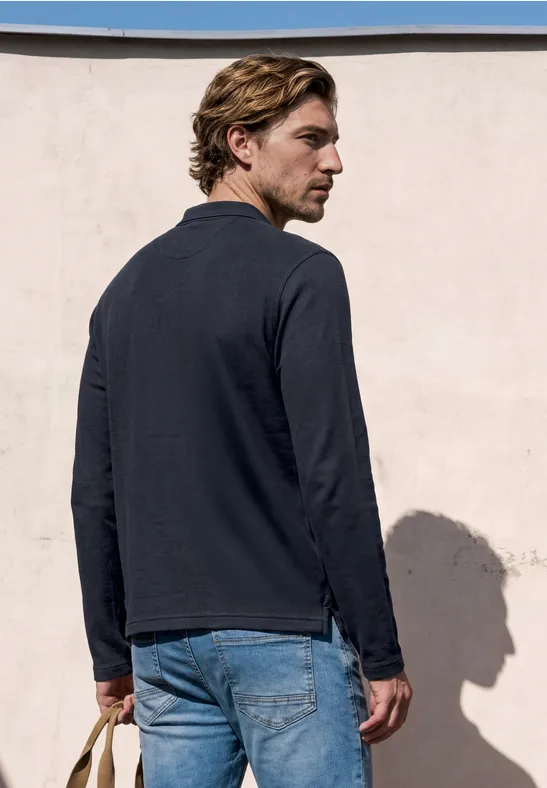 Ein Mann mit blonden Haaren steht vor einer pastellfarbenen Wand, trägt dunkles Oberteil und Jeans.