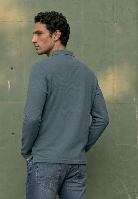 Ein Mann in blauem Polohemd und Jeans steht vor einer grünen Wand und blickt nach rechts.