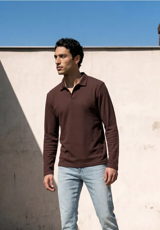 Ein Mann in braunem Langarm-Polo und hellblauen Jeans vor einer hellen Wand
