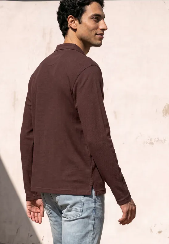 Ein Mann im Profil trägt braunes Hemd und Jeans vor einer hellen Wand.