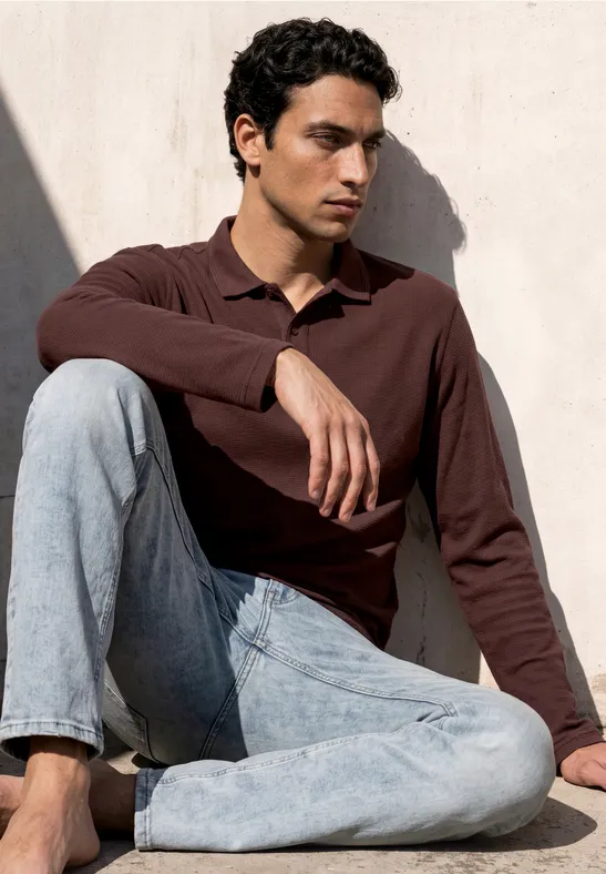 Ein Mann sitzt im Lotussitz vor einer hellen Wand, trägt bordeauxfarbenes Poloshirt und helle Jeans.