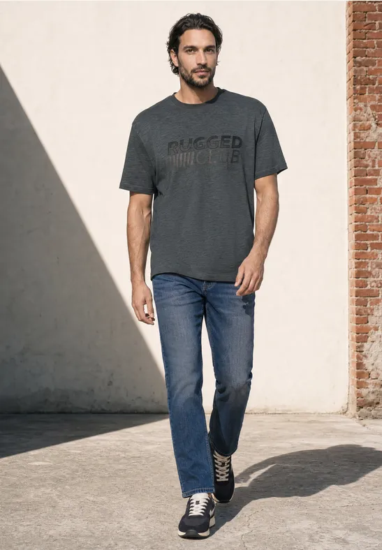 Mann in T-Shirt und Jeans geht draußen auf hellem Hintergrund zu.