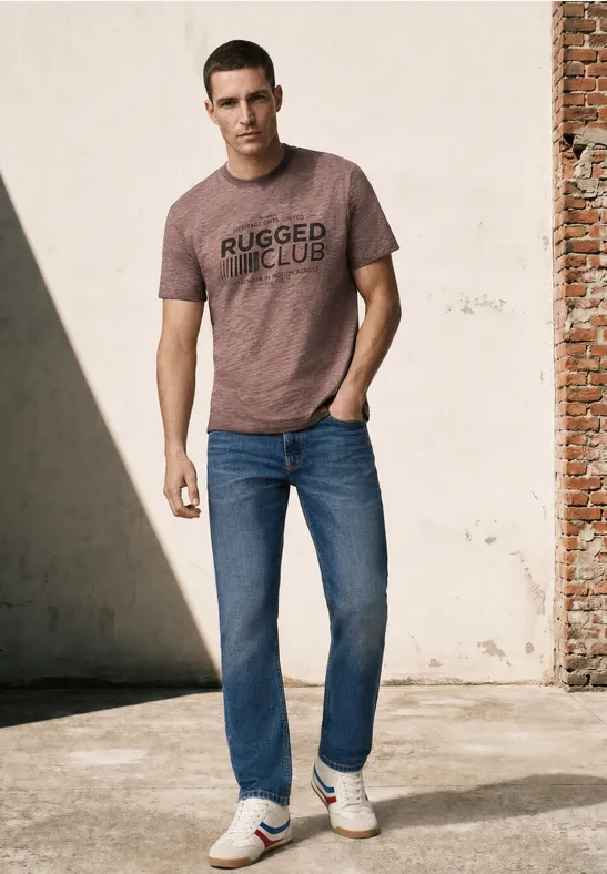 Mann in braunem T-Shirt und Jeans lehnt lässig an einer Wand