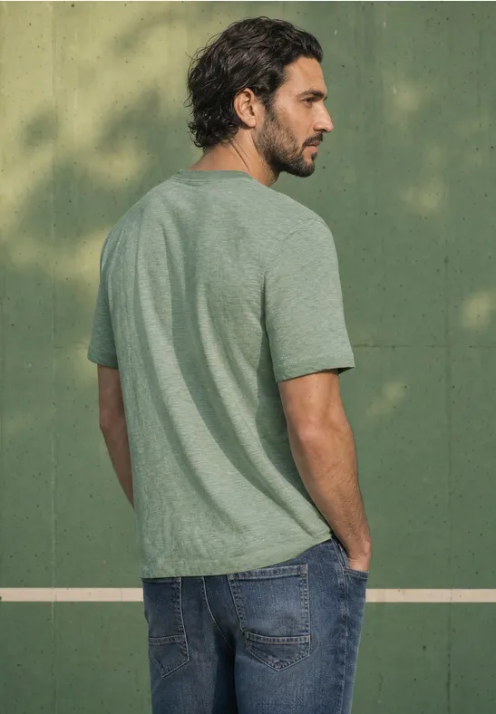 Ein Mann mit braunem Haar trägt ein grünes T-Shirt und Jeans vor einer olivgrünen Wand.