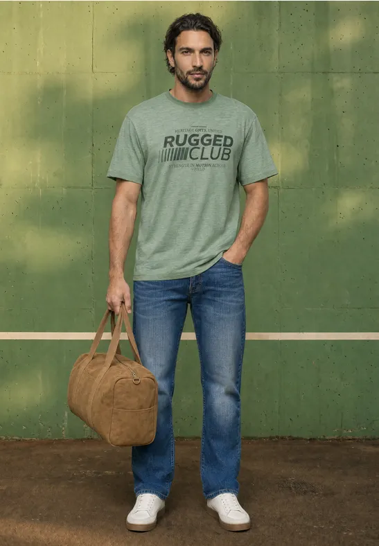 Ein Mann in Jeans und grauem T-Shirt hält eine braune Reisetasche vor einer grünen Wand.