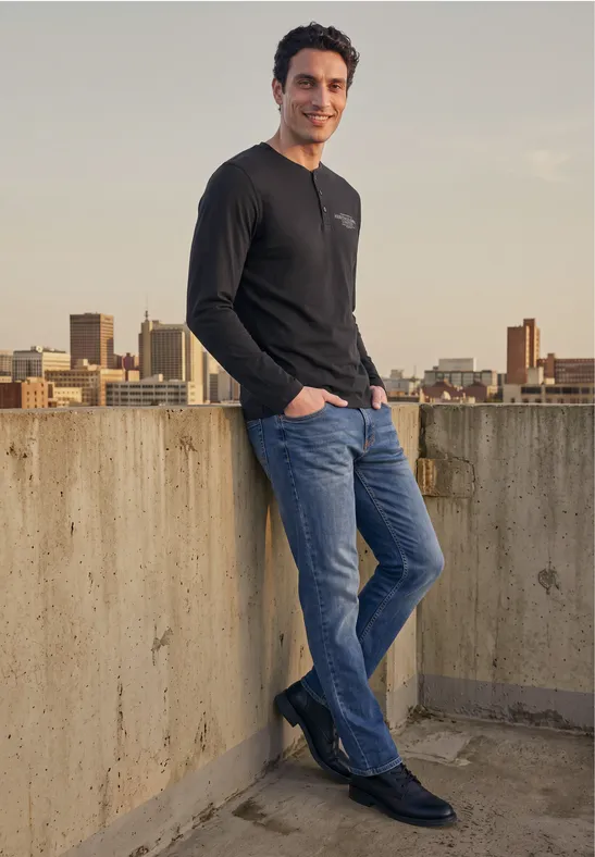 Mann lehnt auf Dachkante, trägt Jeans und schwarzen Pullover, Stadt-Skyline im Hintergrund.