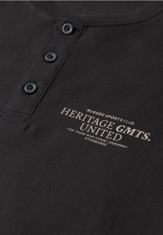 Schwarzes Shirt mit Knopfleiste und goldfarbenem Schriftzug 'Heritage GMTS UNITED'.