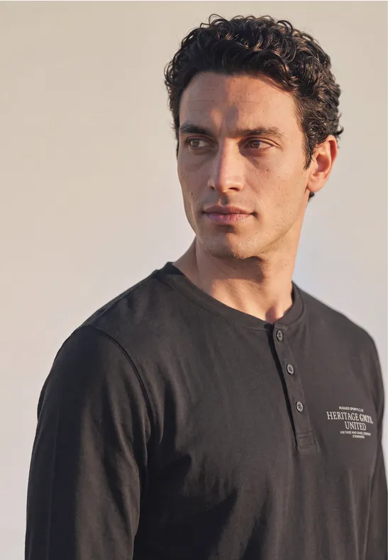 Ein dunkelhaariger Mann in schwarzem Henley-Shirt blickt nach links vor hellem Hintergrund.