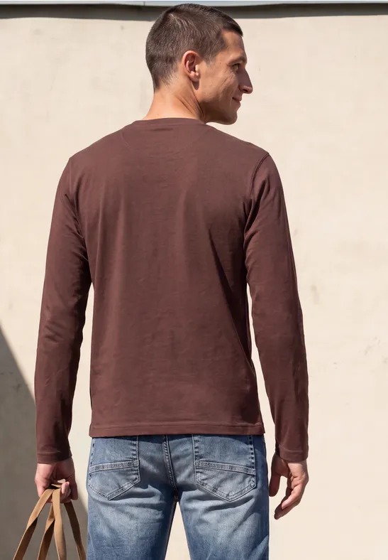 Ein Mann von hinten in braunem Langarmshirt und Jeans, mit gelber Tasche in der Hand vor heller Wand.