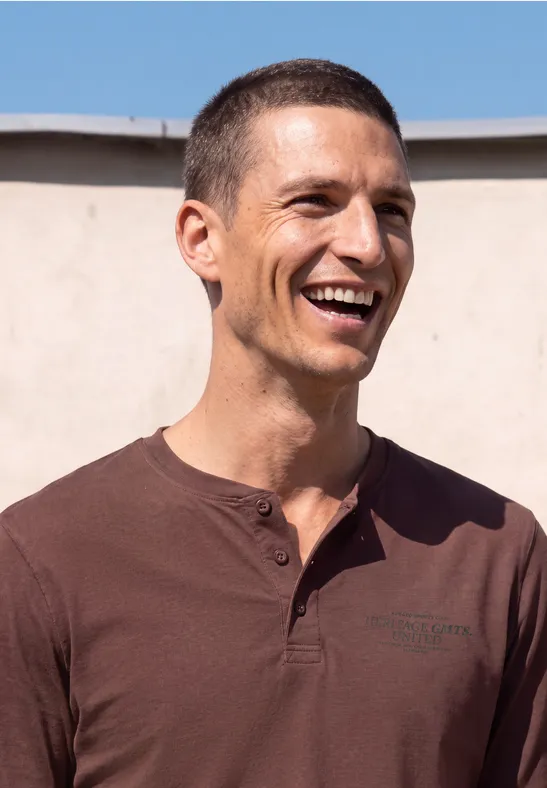 Ein lächelnder Mann mit kurzem dunklem Haar und braunem Shirt vor hellem Hintergrund.